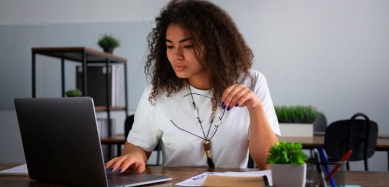 Mulher em um emprego sem ensino superior, diante do notebook com o óculos na outra mão