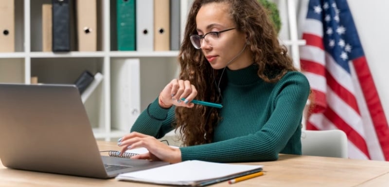 Mulher com notebook, caderno aberto, a bandeira dos Estados Unidos atrás dela