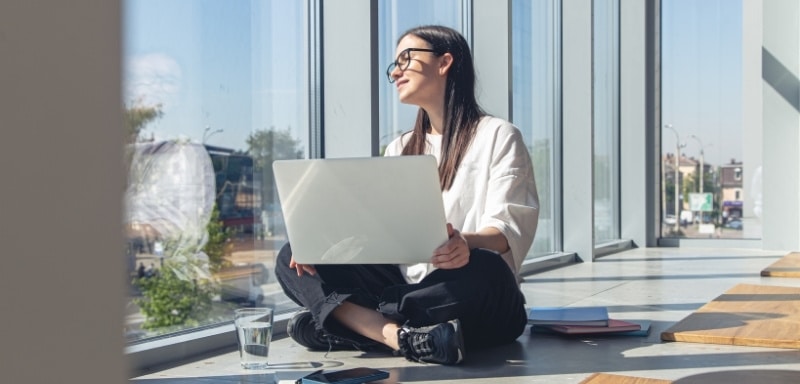 Mulher com um notebook em mãos, sentada diante de uma janela com o céu azul e ensolarado